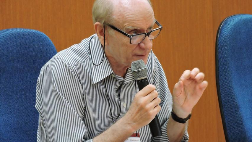 Foto do professor Michel Thiollent em uma palestra, falando ao microfone.